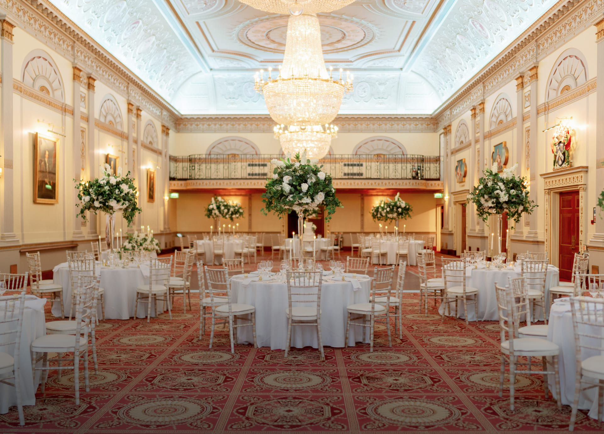 The Great Hall laid with round tables and white chairs under crystal chandeliers, with tall floral centrepieces and a gallery at the far end, Plaisterers' Hall