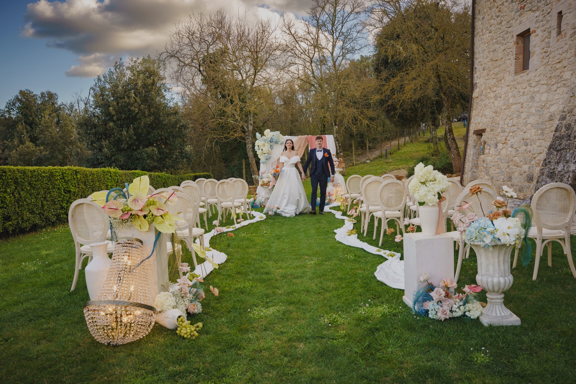 Couple at the end of a lawn ceremony aisle lined with floral urns and white chairs, beside the stone walls, Relais La Costa