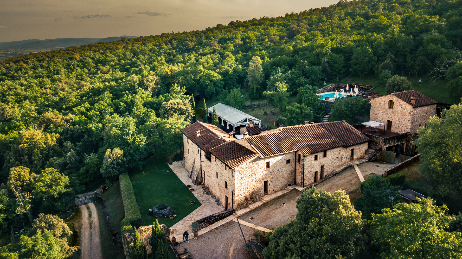 Aerial view of the stone buildings surrounded by woodland, with the pool and a white marquee, Relais La Costa