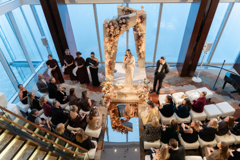 A wedding ceremony seen from the staircase, with guests seated either side of a floral arch in front of the windows, Shangri-La The Shard, London