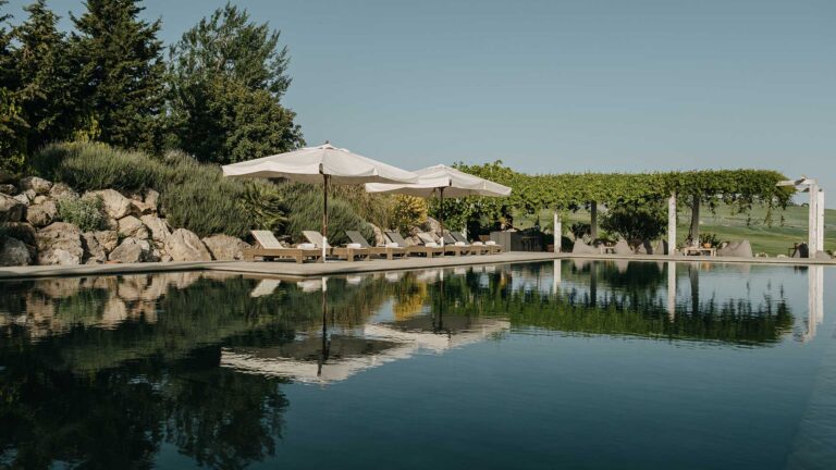 The pool reflecting white parasols and loungers, with a pergola and rock garden behind, Susafa