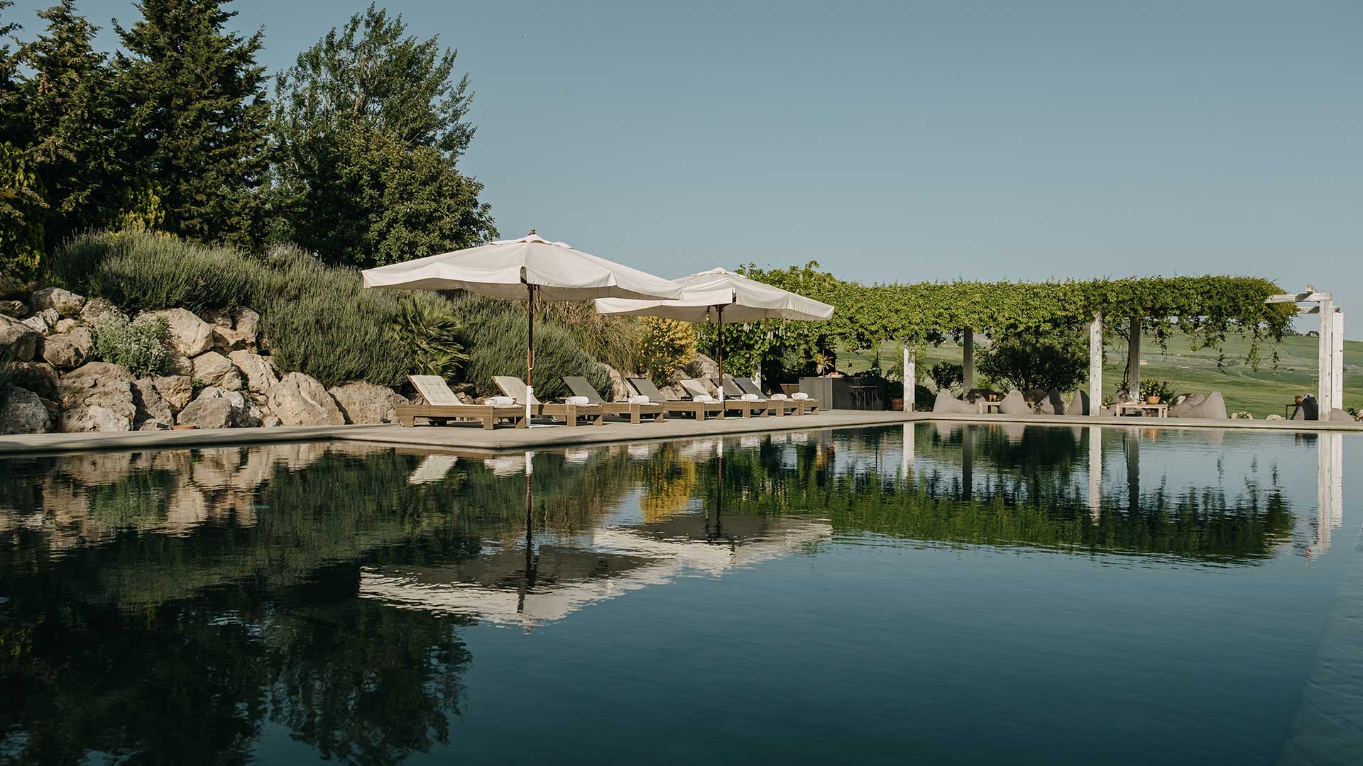 The pool reflecting white parasols and loungers, with a pergola and rock garden behind, Susafa
