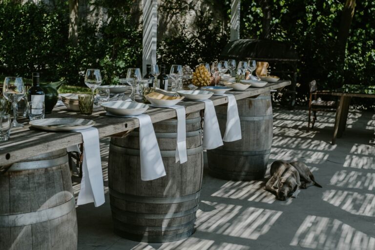 A long table laid on wine barrels under a pergola, with dappled light on the paving, Susafa