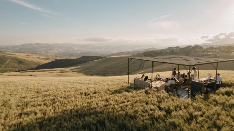 Guests at tables under an open steel pavilion in the wheat fields, with hills beyond, Susafa