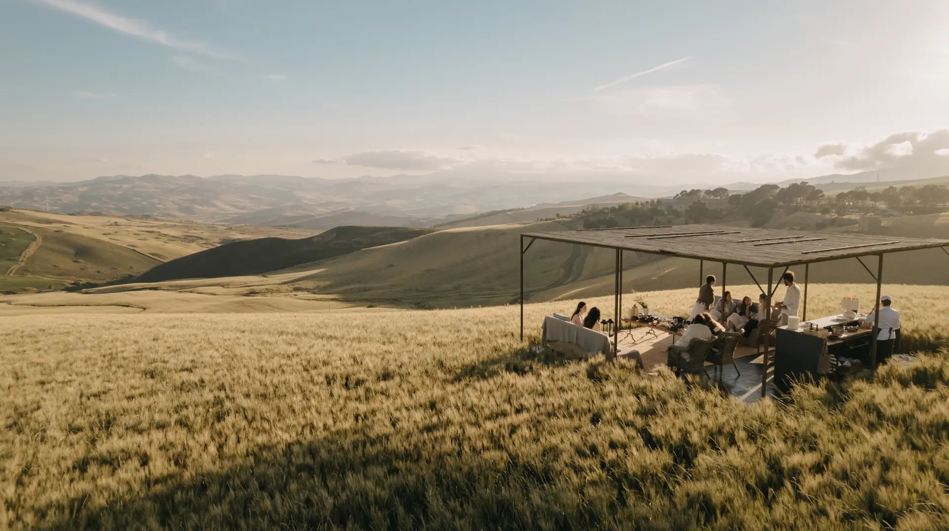 Guests at tables under an open steel pavilion in the wheat fields, with hills beyond, Susafa