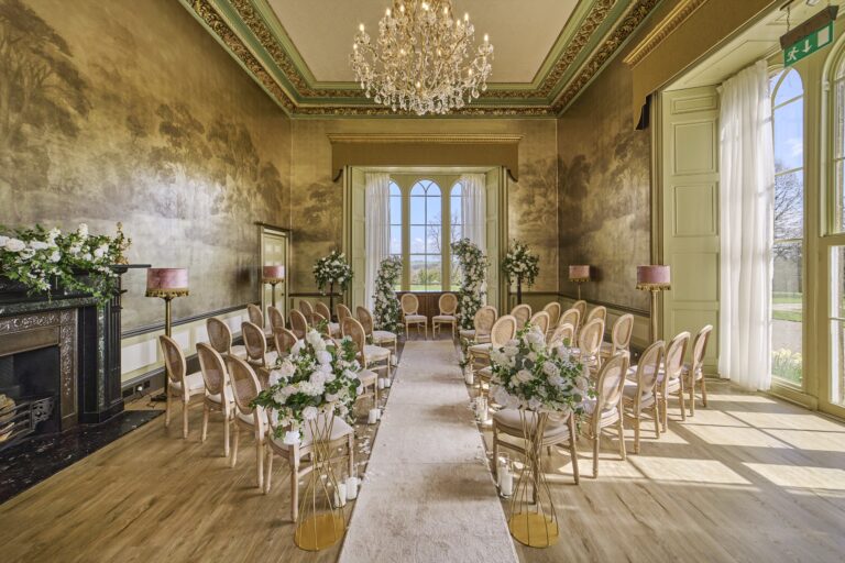 The Ballroom set for a ceremony, with rows of cane chairs, white flowers and tall arched windows, Swinton Park