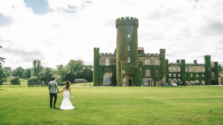 A bride and groom, seen from behind, walking hand in hand across the lawn towards the castle, Swinton Park