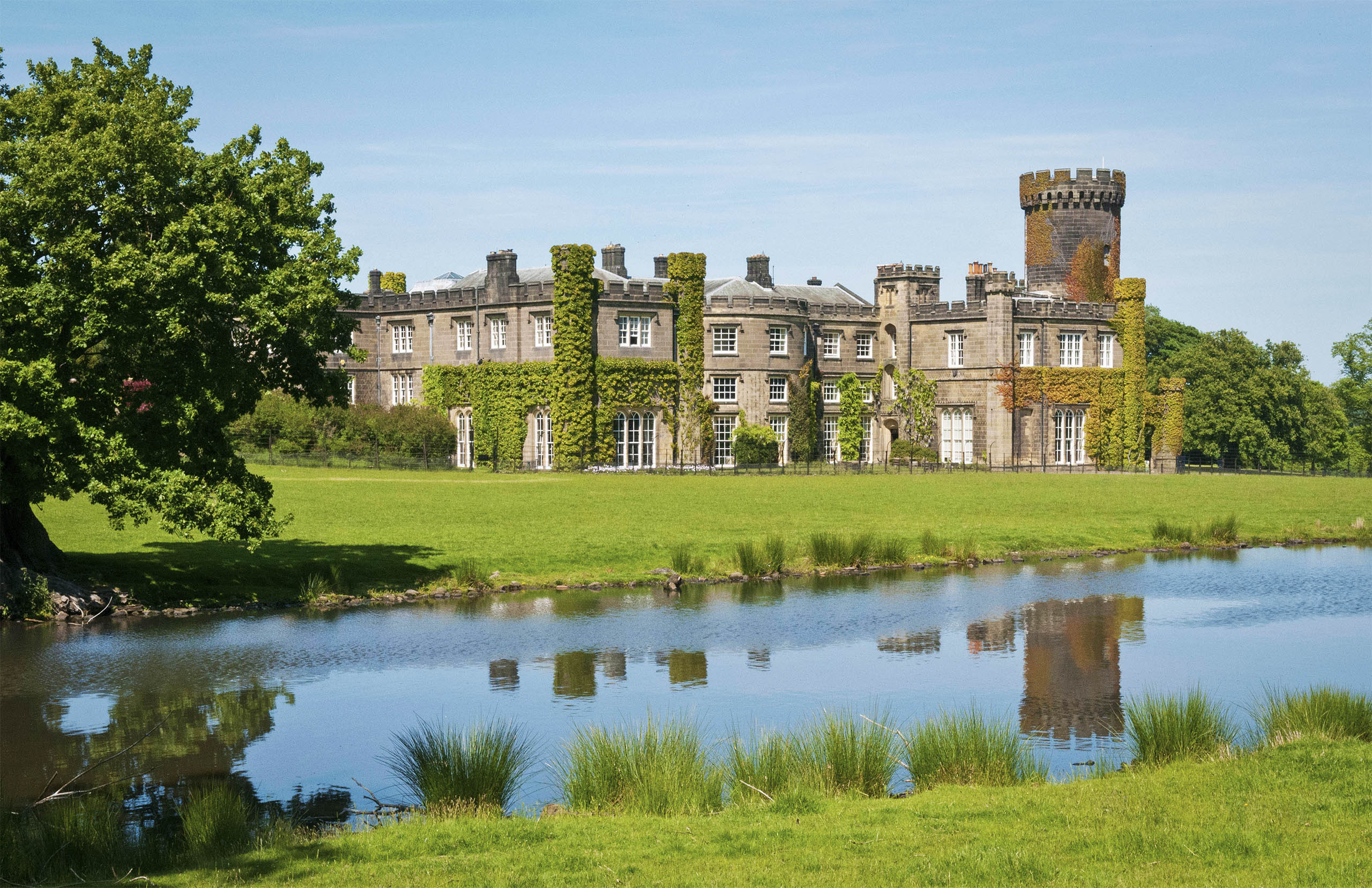 The castle front seen across Home Lake, with the round tower at the east end and ivy on the walls, Swinton Park