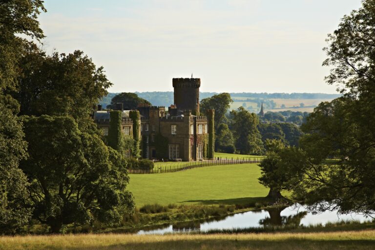 The castle and tower framed by mature trees above a stretch of water in the parkland, Swinton Park