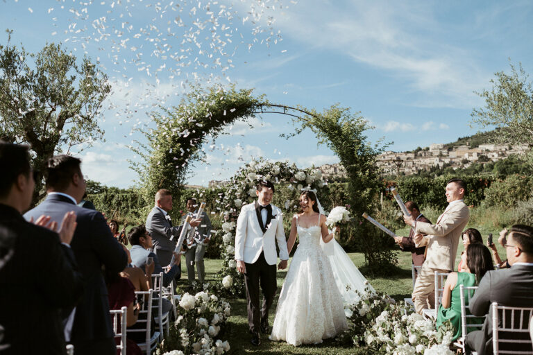 A couple walking back down the aisle under a flowered arch in the garden, guests either side, Tenuta San Masseo