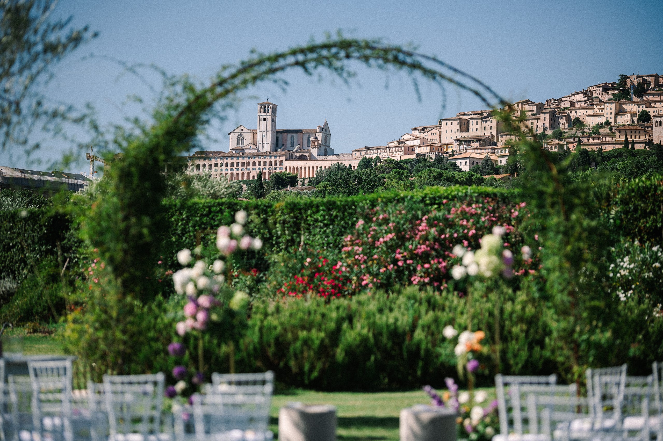 Ceremony chairs and a flowered arch in the garden, framing the Basilica of Saint Francis, Tenuta San Masseo