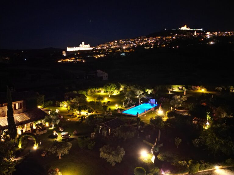 Night drone view of the lit gardens and pool, with Assisi illuminated on the hill, Tenuta San Masseo