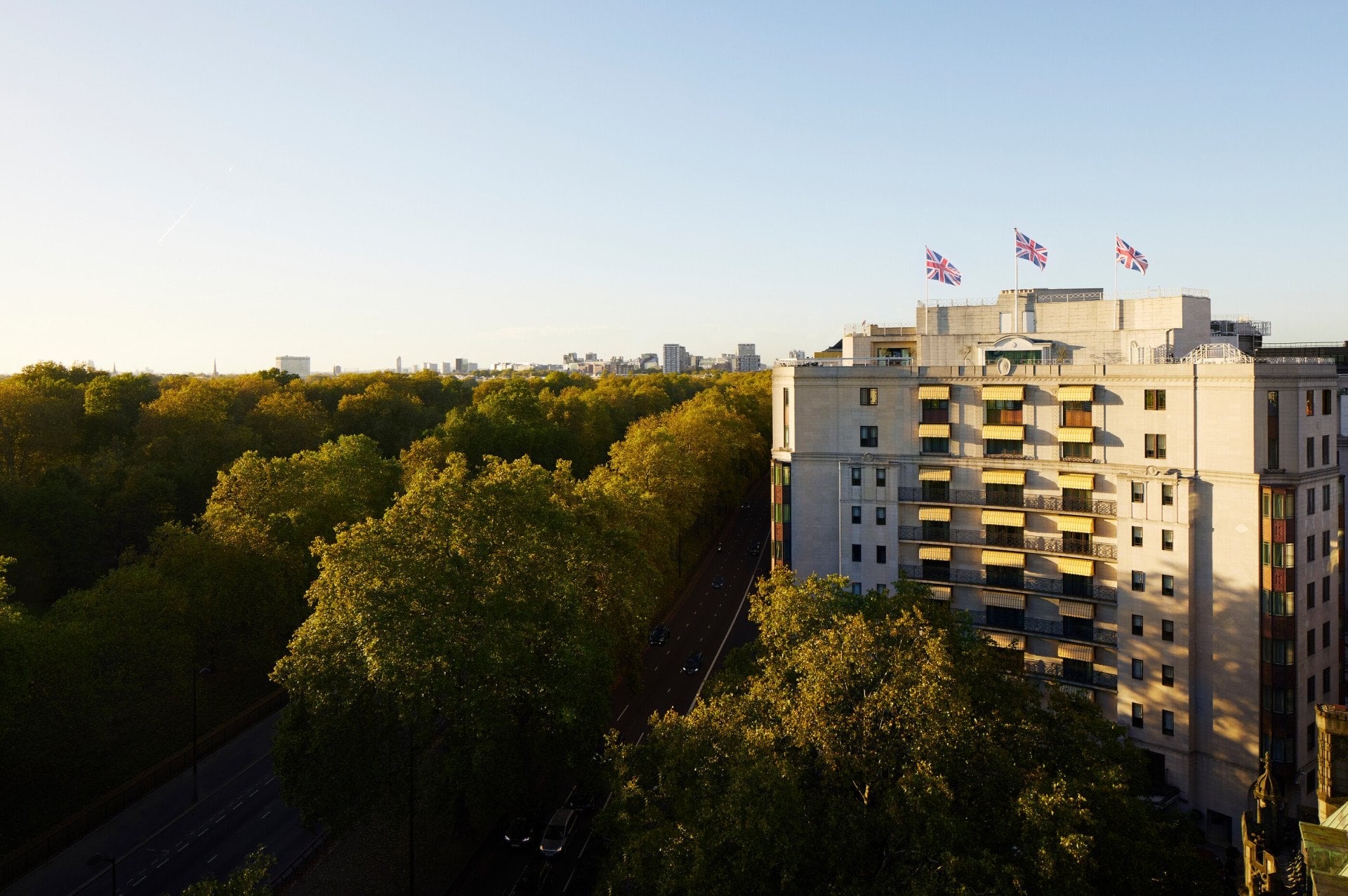 The hotel's white facade and rooftop flags above the trees of Hyde Park in early evening light, The Dorchester