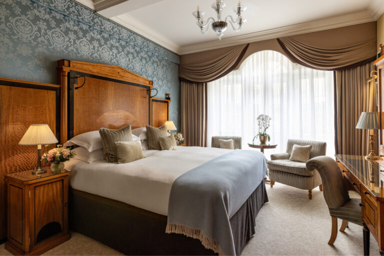 A bedroom with a wooden headboard, a pale blue throw and two armchairs by the window, The Goring