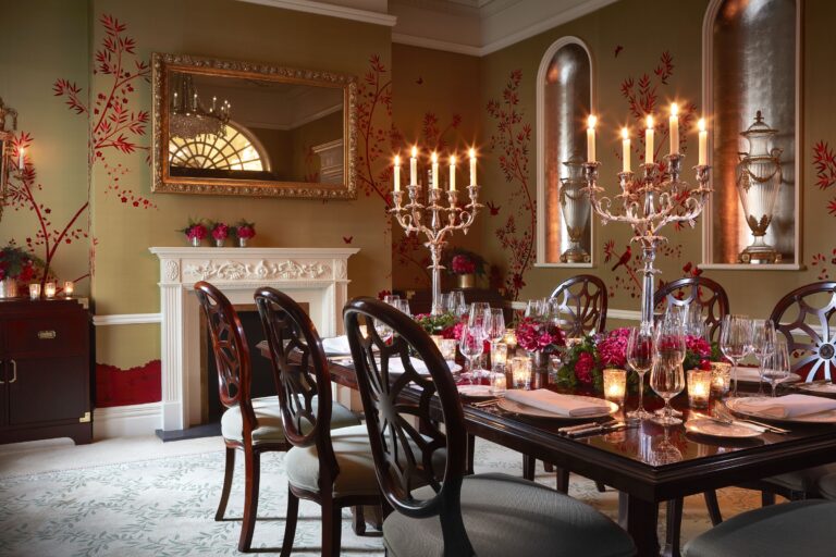 A candlelit table with a silver candelabra in a room with red floral silk walls and a marble fireplace, The Goring