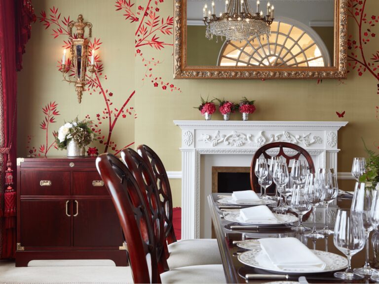 A laid dining table by a white fireplace, with red flowers on the mantel and a gilt mirror above, The Goring