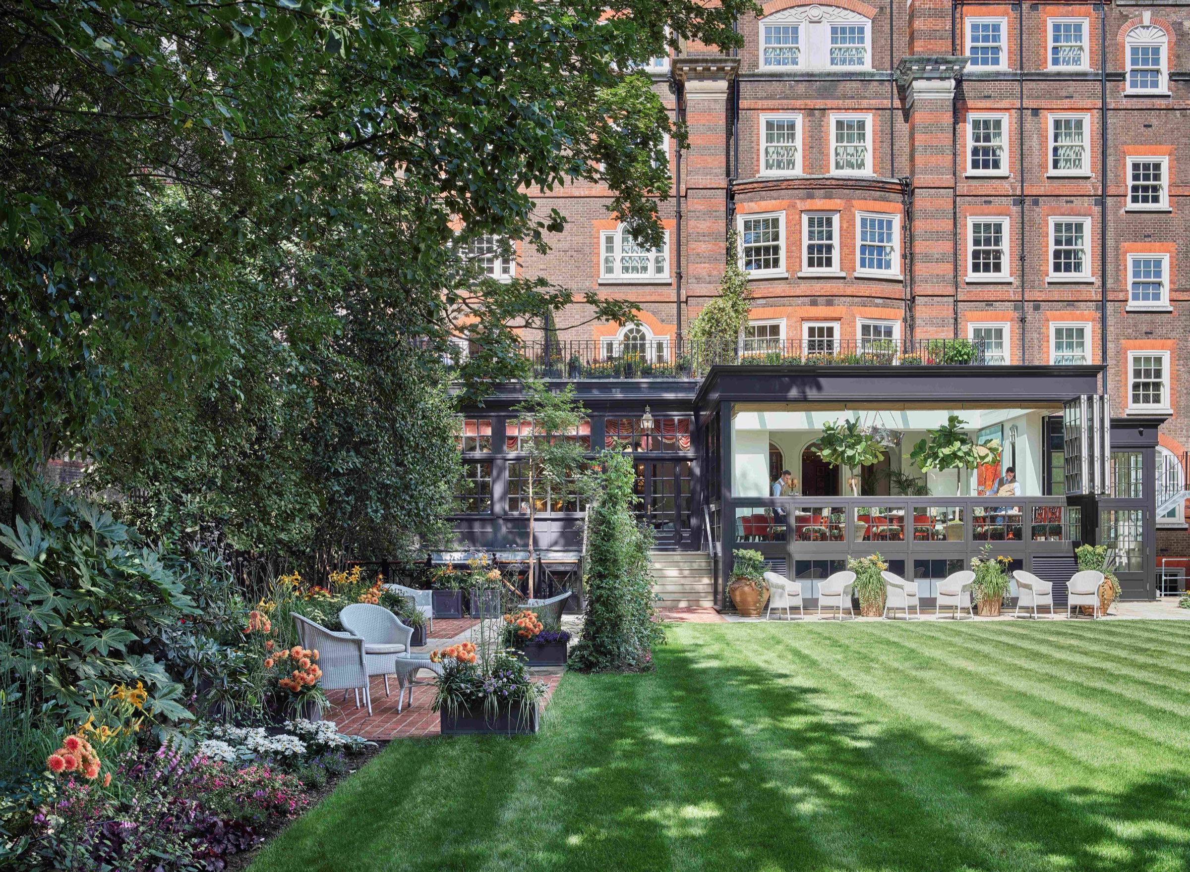 The long striped lawn of the private garden, with borders, trees and the red-brick back of the hotel beyond, The Goring