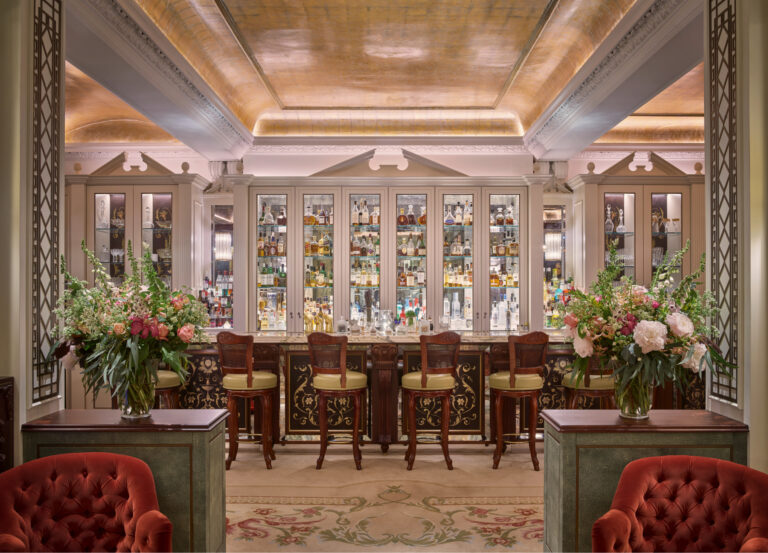 The bar with glass-fronted cabinets of bottles, high stools and flowers along the counter, The Goring