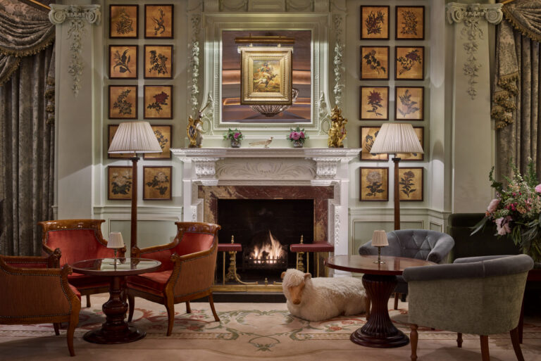 A lounge with a lit fireplace, armchairs and walls hung with framed botanical prints, The Goring