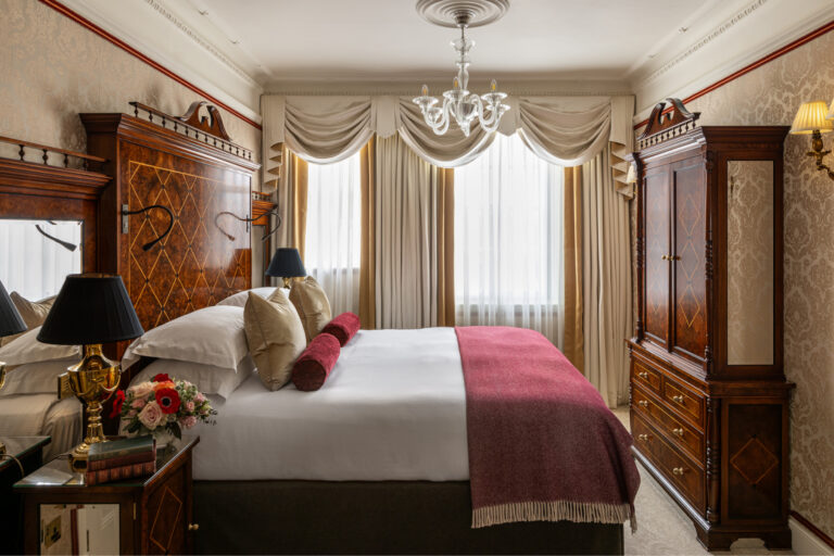A bedroom with a tall marquetry headboard, matching wardrobe and a burgundy throw, The Goring