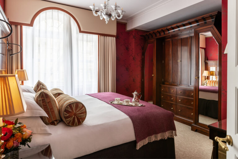 A bedroom with red silk walls, an arched window, a wooden wardrobe and bolster cushions, The Goring