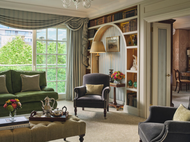 A sitting room with a green sofa, a bookcase alcove and tall windows onto trees, The Goring