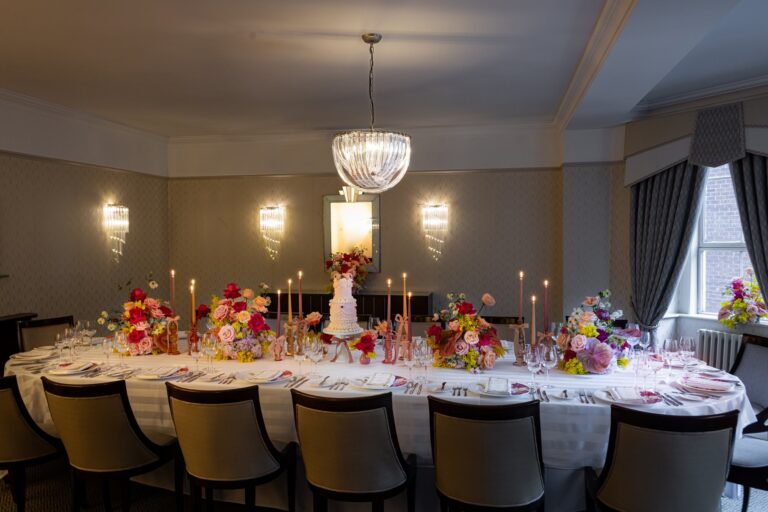 A long candlelit table with pink and red flowers under a chandelier in the Findlay Room, The Lansdowne Club
