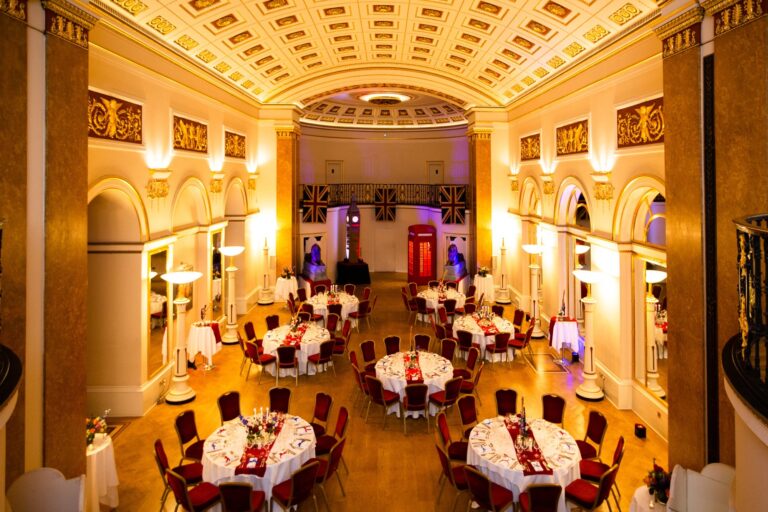 The Ballroom laid for a large dinner with red chairs and a stage at the far end, The Lansdowne Club