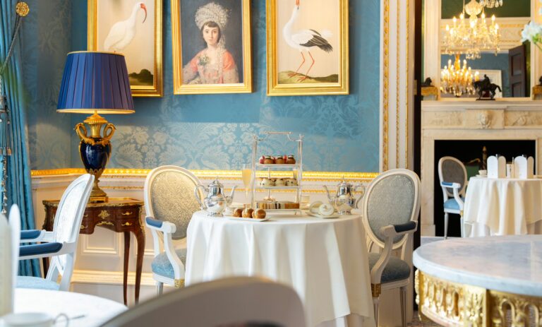 A tiered afternoon tea stand on a round table in a blue room with paintings, The Ritz London