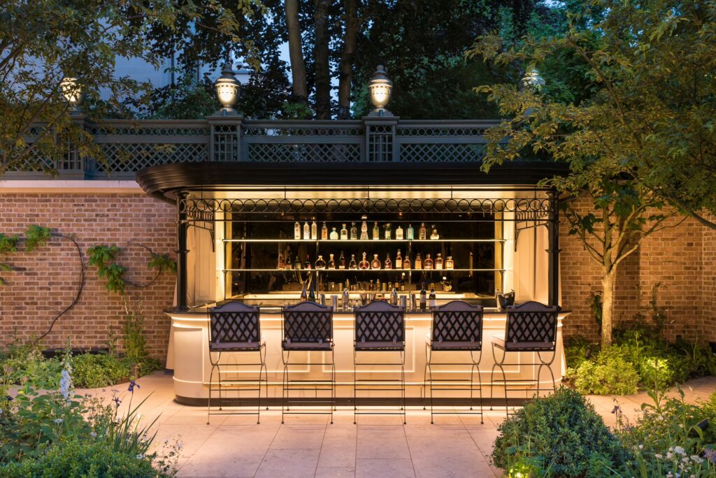 The Secret Garden bar lit at dusk, with stools in front of a bottle-lined counter, The Ritz London