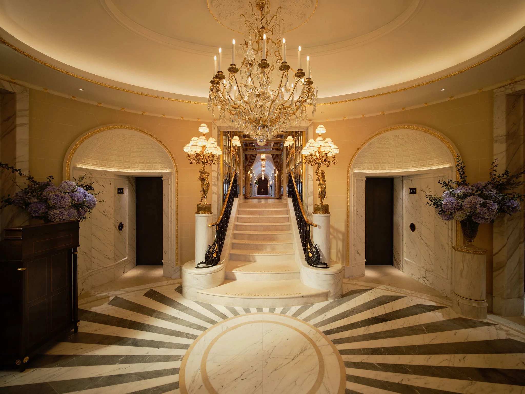 An oval hall with a marble floor, a chandelier and a staircase rising between two archways, The Ritz London