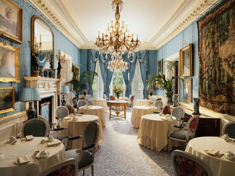 The Queen Elizabeth Room laid with round tables, under chandeliers, with blue walls and a tapestry, The Ritz London