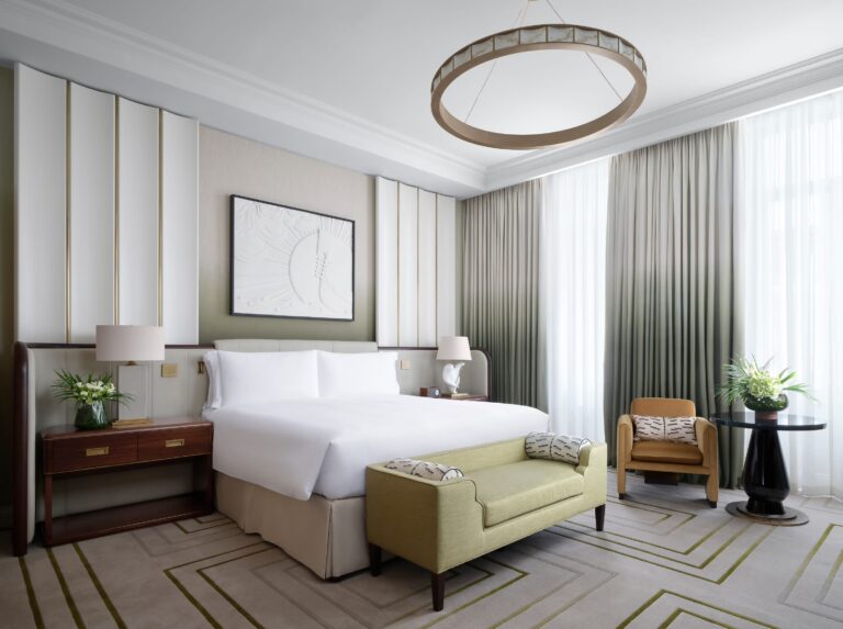 A bedroom with a king bed, a pale sofa at its foot and a patterned carpet, The Savoy