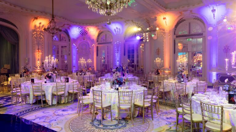 Round tables laid for dinner in the Lancaster Ballroom under coloured lighting, The Savoy