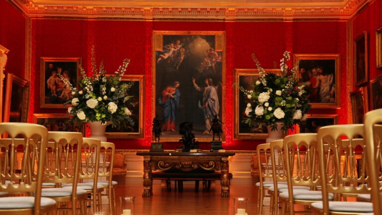 Gilt chairs set out for a ceremony in a red-walled gallery, with tall white flower arrangements either side of a large painting, The Wallace Collection