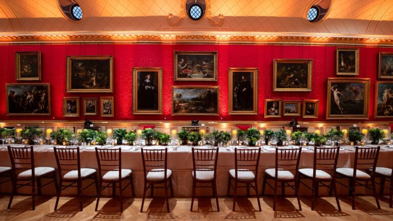 A long dinner table with greenery and candles beneath paintings on red walls, The Wallace Collection