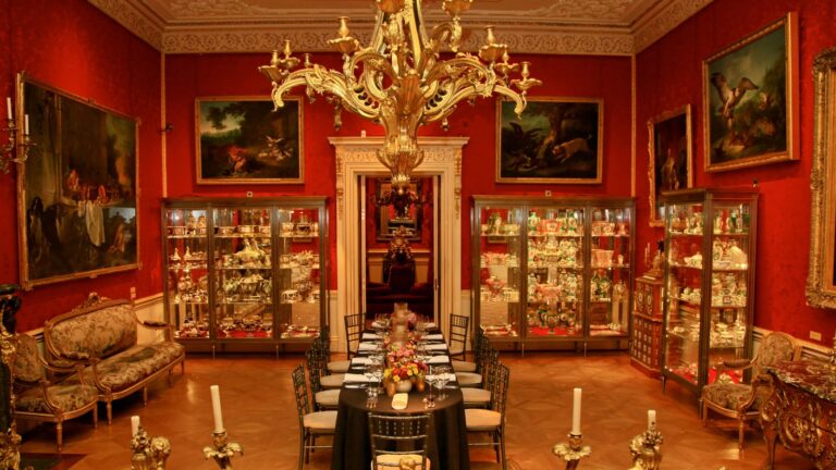 A red-walled room with a gilt chandelier, glass display cabinets and a table laid for dinner, The Wallace Collection