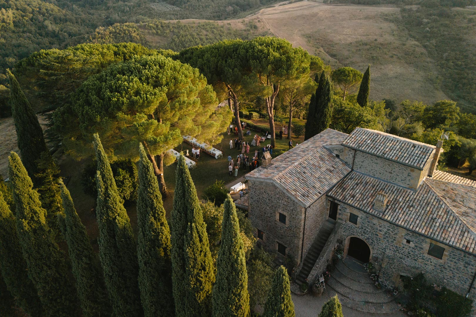 Aerial view of the stone tower house among cypresses and umbrella pines, with guests gathered on the lawn, Torre Collevento