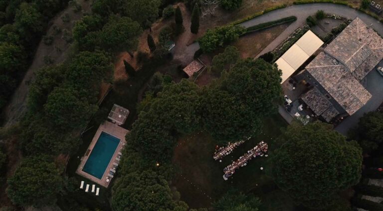 Overhead view of the estate at dusk: the tower roofs, a white canopy, long dinner tables in the garden and the pool below, Torre Collevento