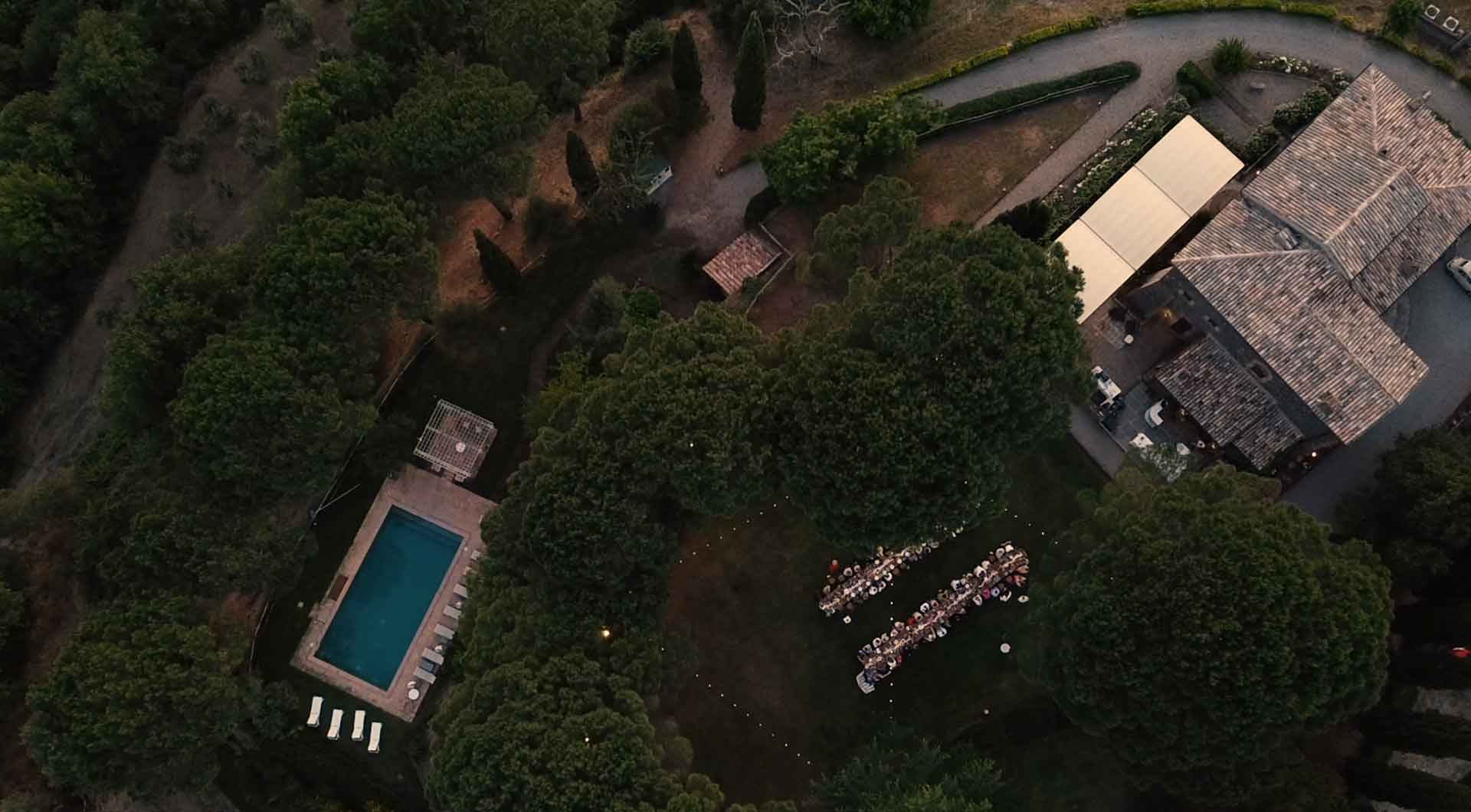 Overhead view of the estate at dusk: the tower roofs, a white canopy, long dinner tables in the garden and the pool below, Torre Collevento