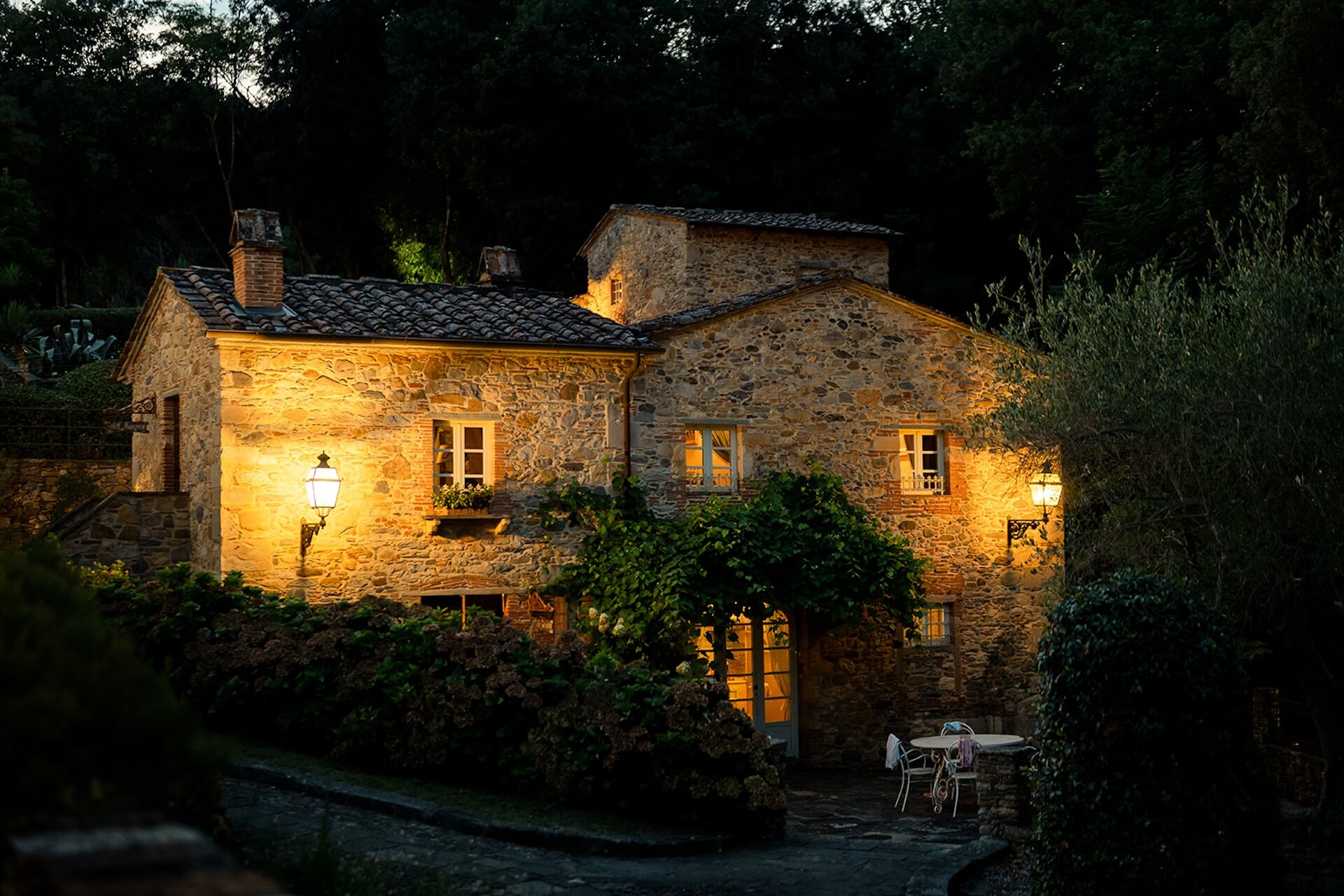 A stone farmhouse in the borgo lit at dusk, with a table under a pergola, Valle di Badia