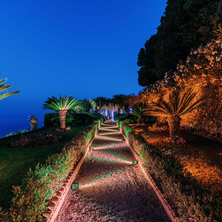 A lit garden path through palms and shrubs at night, Villa La Rondinaia