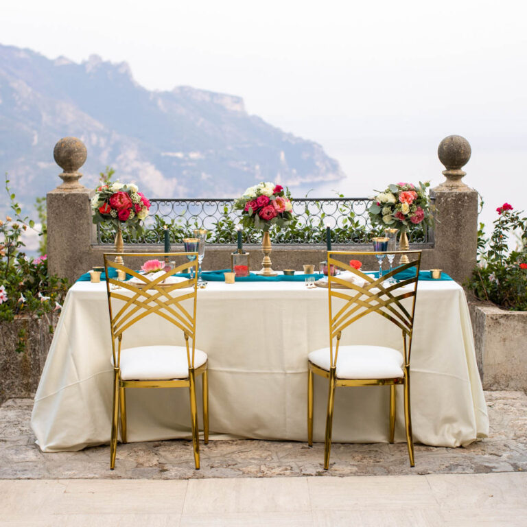 A table for two with gold chairs on a terrace, the coast behind, Villa La Rondinaia
