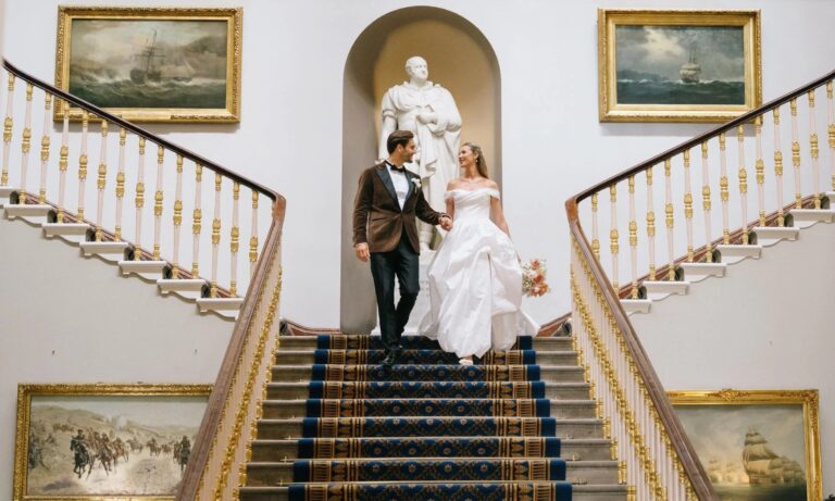 The couple descending the Grand Staircase beneath the marble statue niche, gilt-framed marine paintings either side, 116 Pall Mall