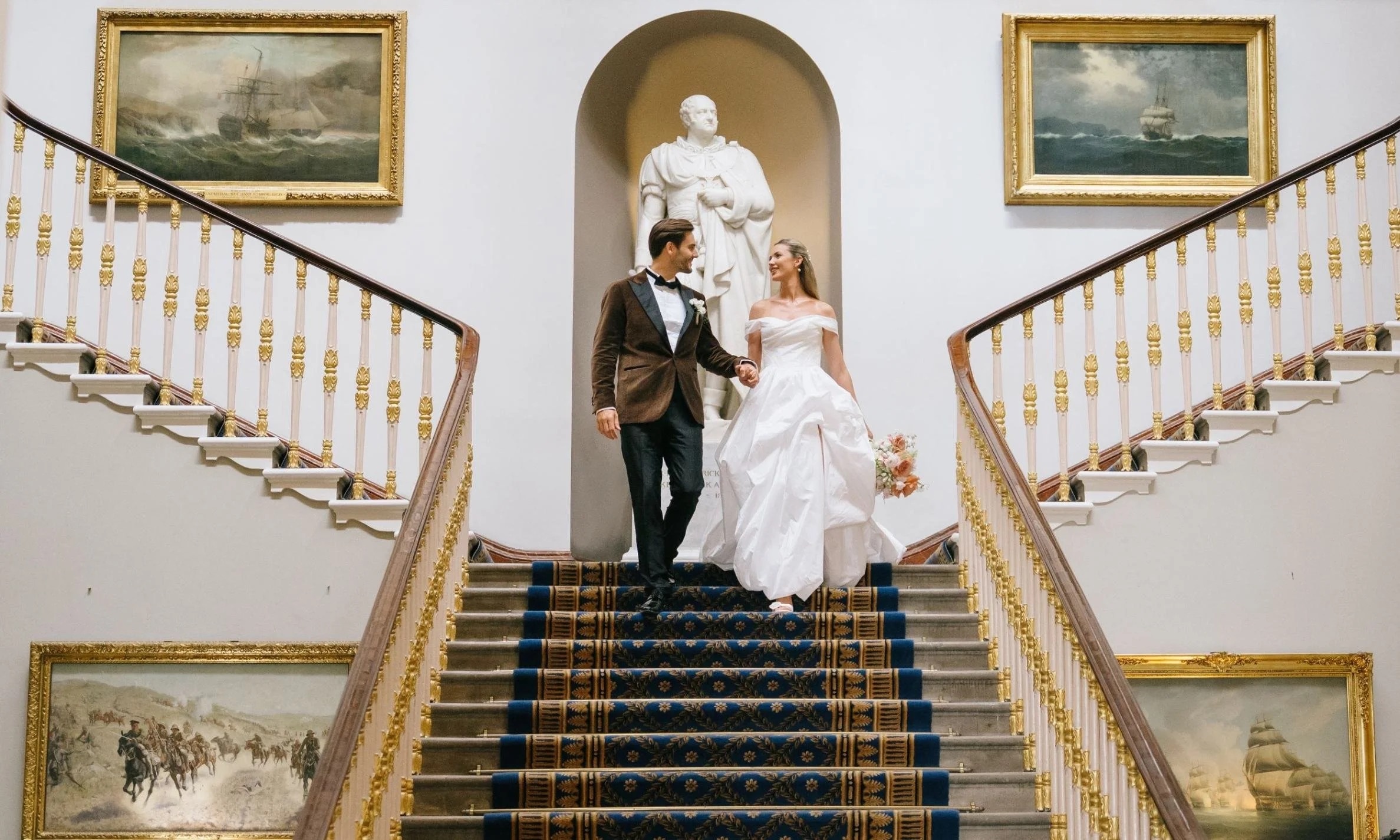 The couple descending the Grand Staircase beneath the marble statue niche, gilt-framed marine paintings either side, 116 Pall Mall