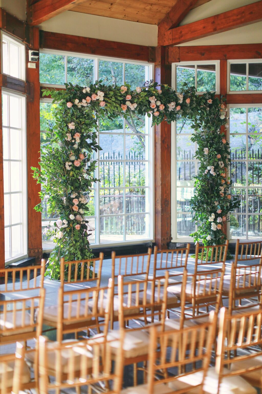 Floral ceremony arch and wooden chairs in the glass pavilion at the Central Park Boathouse, 5th Avenue Weddings