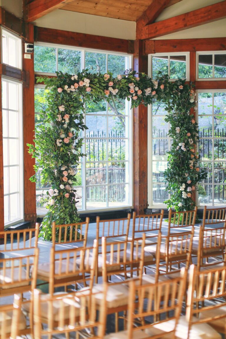 Floral ceremony arch and wooden chairs in the glass pavilion at the Central Park Boathouse, 5th Avenue Weddings