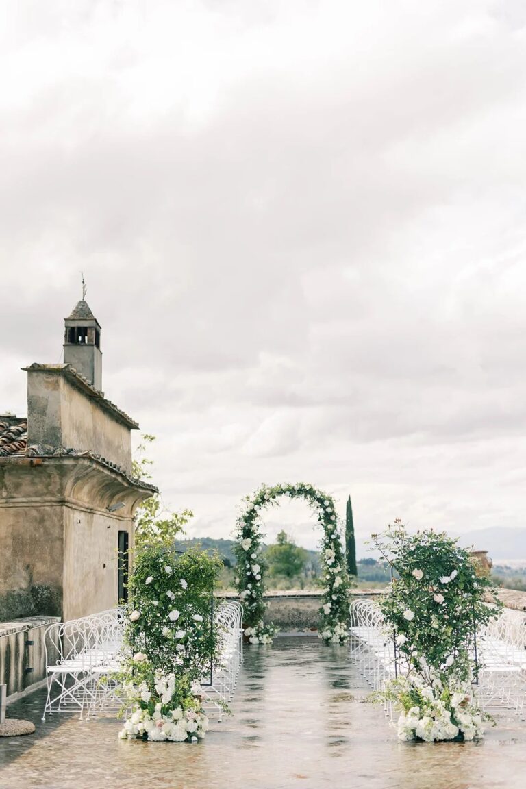 Floral ceremony arch in a Tuscan courtyard beside a small chapel, 5th Avenue Weddings