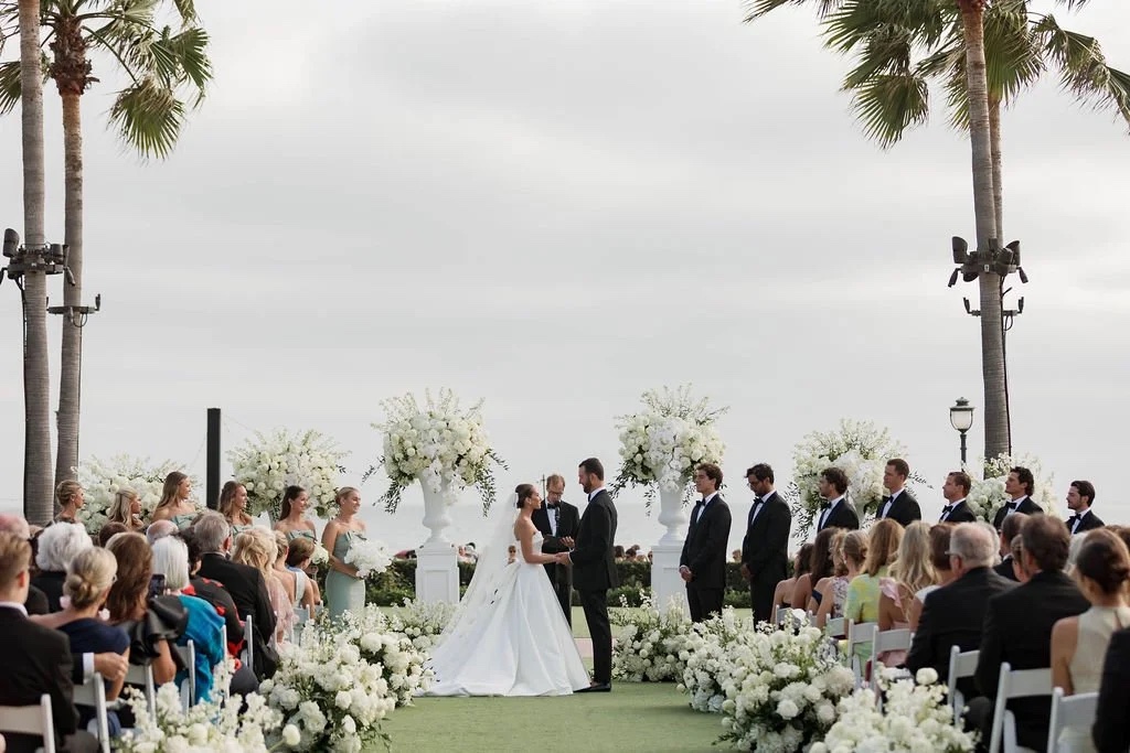 An oceanfront lawn ceremony between white flower arrangements