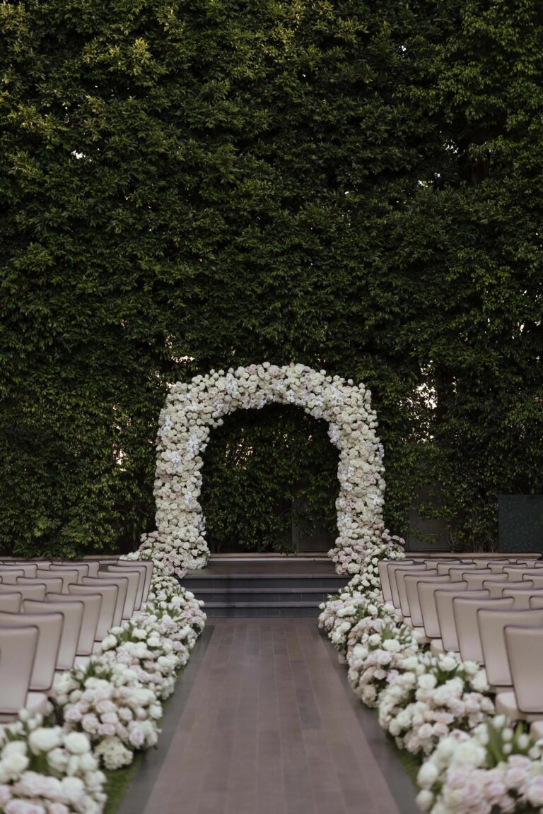 White floral arch at the end of an aisle lined with white flowers against a hedge wall, About Details Details